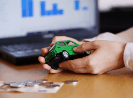 Foto mostrando as maos de uma pessoa segurando um carro em miniatura verde, com cédulas e moedas na mesa. ao fundo a tela de um notebook mostrando gráficos