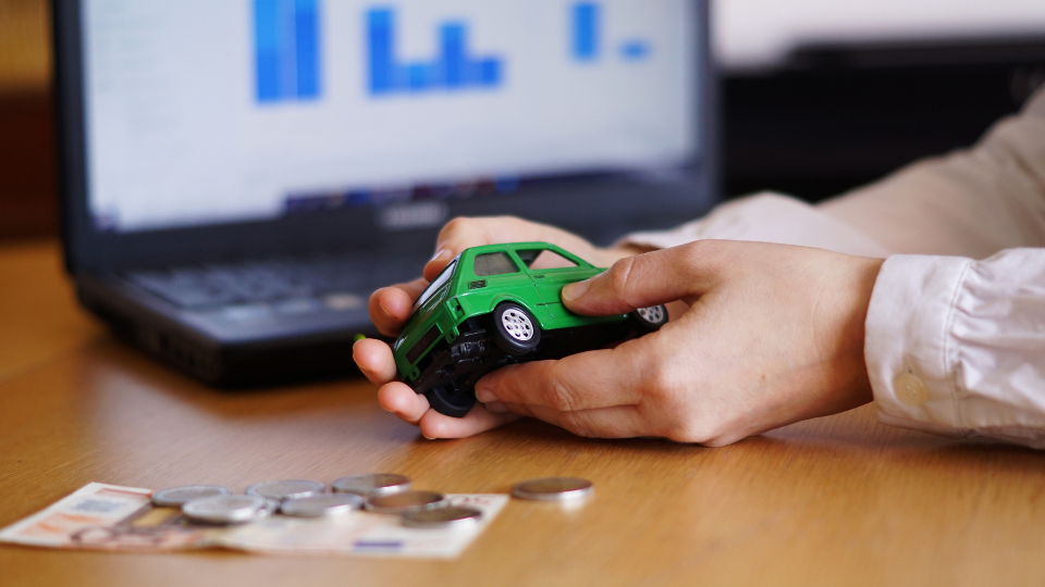 Foto mostrando as maos de uma pessoa segurando um carro em miniatura verde, com cédulas e moedas na mesa. ao fundo a tela de um notebook mostrando gráficos