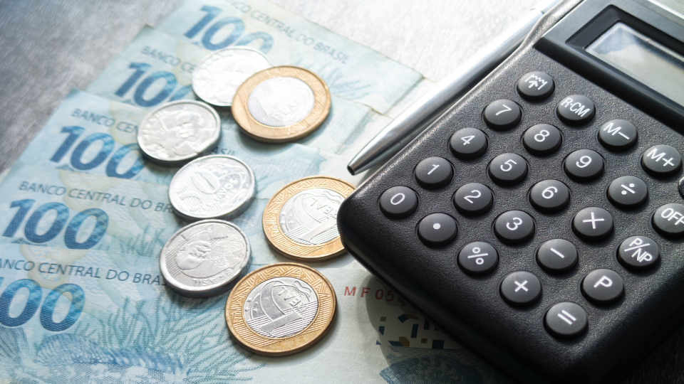 calculadora e reais brasileiros na mesa com cédulas e moedas junto de uma caneta prata