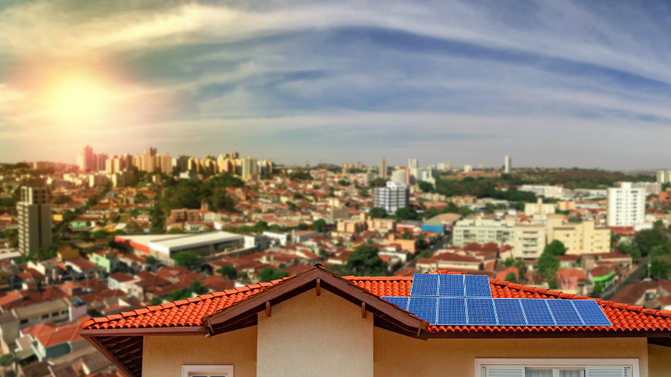 Casa no primeiro plano com placas solares em seu telhado ao fundo uma cidade iluminada por um sol bem brilhoso